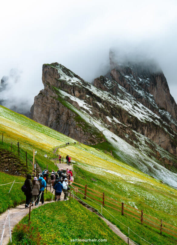 A group of hikers walk along a winding trail on a green hillside with patches of snow, leading toward the dramatic Seceda ridgeline and steep, rocky mountains partially covered by clouds.