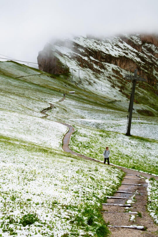 A man walks up a winding path through a grassy, snow-dusted hillside near the Seceda ridgeline, with power lines and low clouds covering a rocky mountain of dolomites, italy