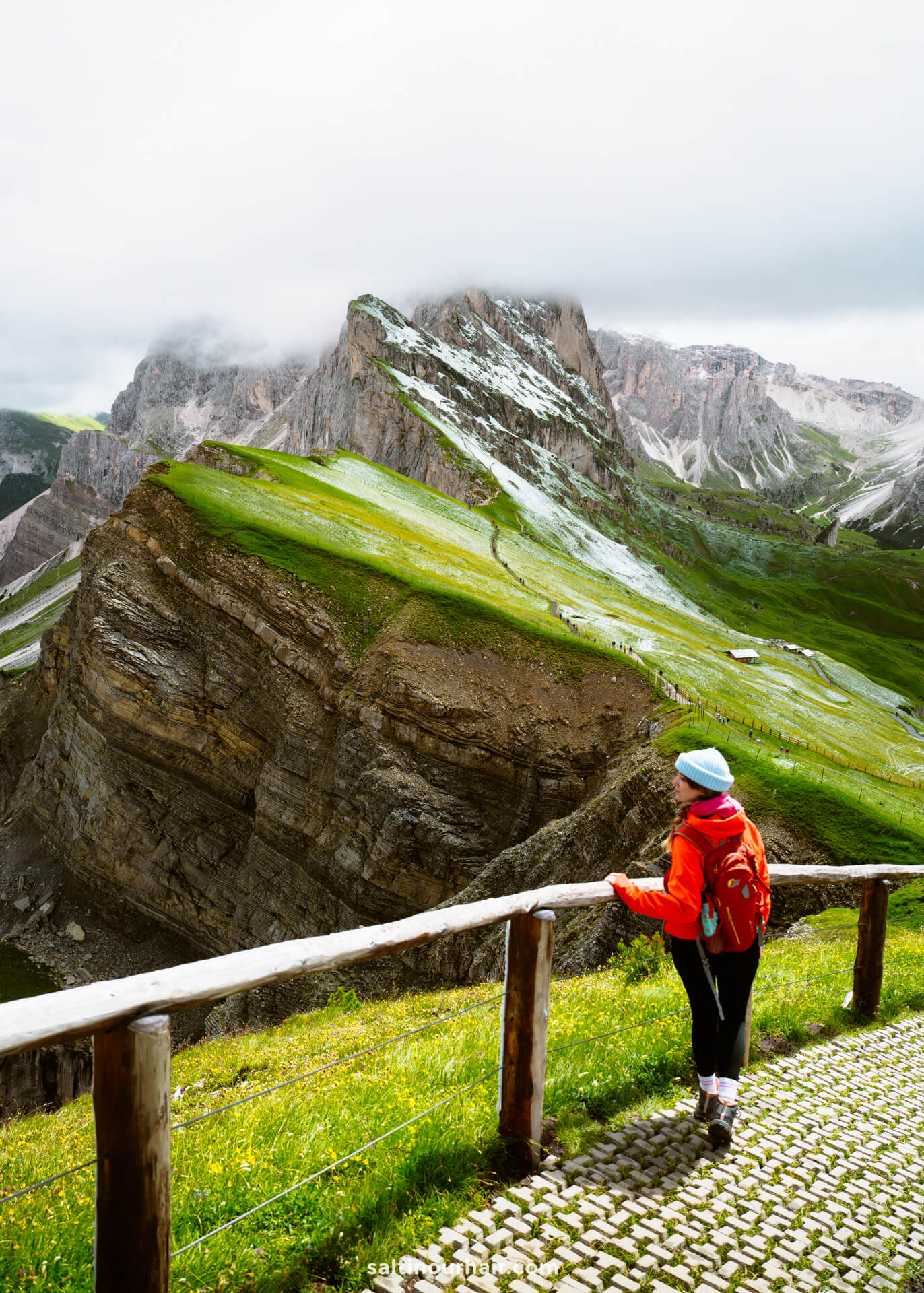 A women in a red jacket and blue hat stands by a wooden railing, gazing at the dramatic green and rocky Seceda ridgeline in the dolomites italy, partly shrouded by clouds.