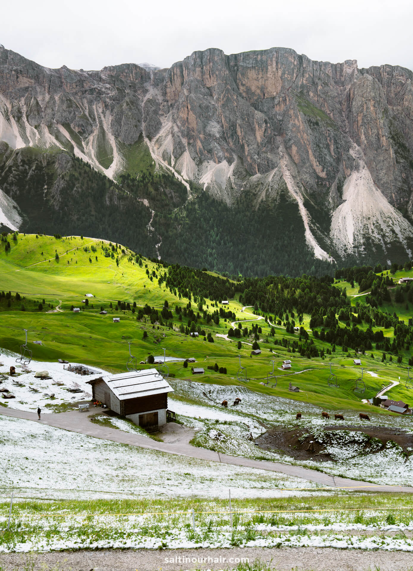A rustic barn sits on a snowy patch amid green fields near the Seceda ridgeline in the dolomites, with scattered houses and dramatic mountains in the background under a cloudy sky.