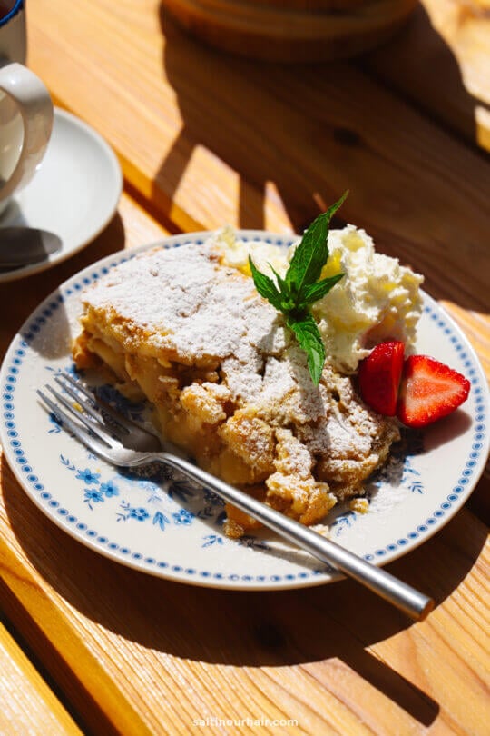 A plate with a slice of apple crumble topped with powdered sugar, whipped cream, strawberries, and a mint sprig sits on a wooden table with a fork—perfect for savoring after a hike on the scenic Alpe di Siusi.