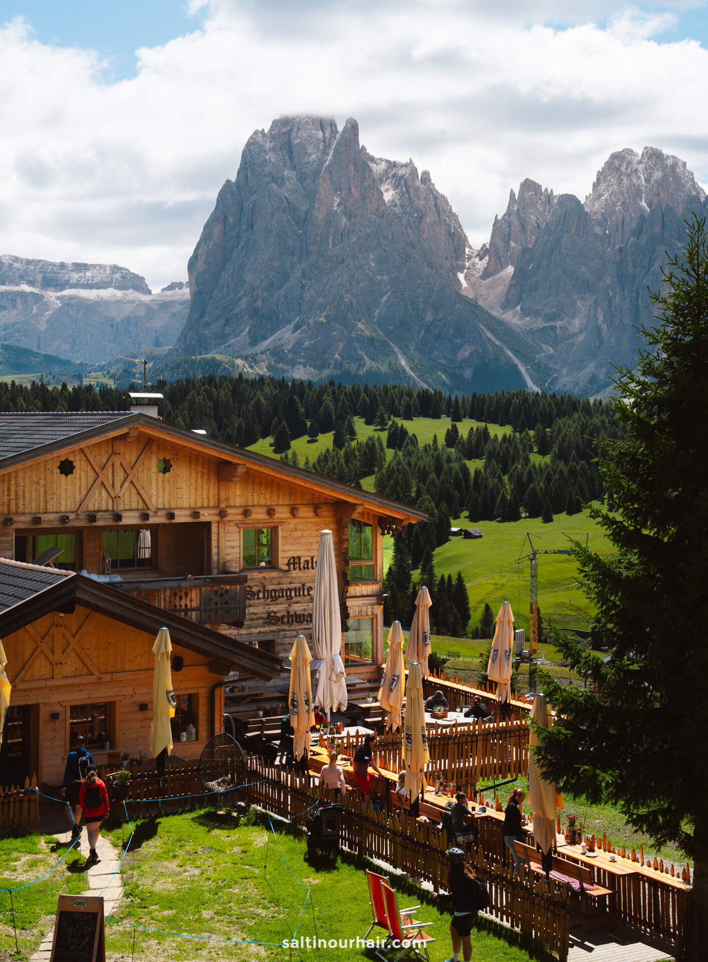 A wooden mountain lodge restaurant with outdoor seating sits in a green valley on Seiser Alm, backed by forested hills and the rugged Dolomites mountains of Italy under a partly cloudy sky.