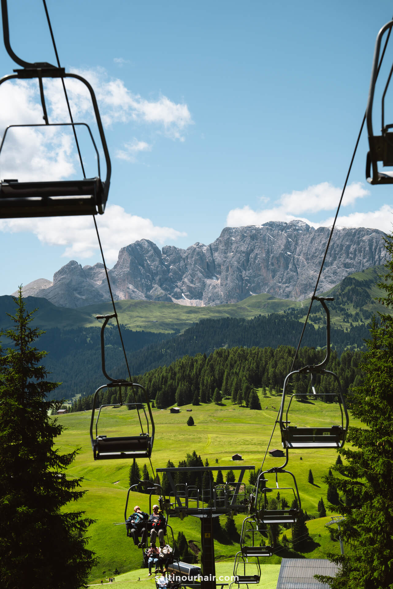 Empty ski lift chairs ascend over the green alpine landscape of Seiser Alm, with trees and distant Dolomites mountains in Italy under a clear blue sky.