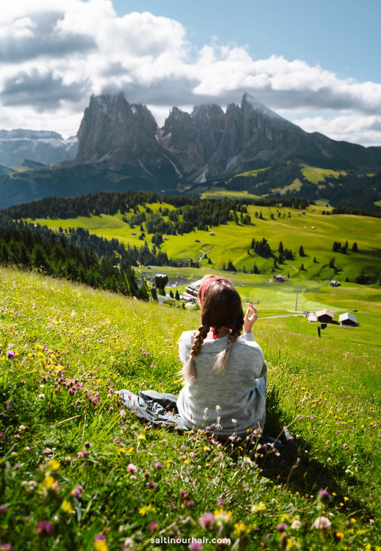 A woman with braided hair sits in a grassy field of wildflowers on the Seiser Alm, facing distant Dolomites mountains and a valley dotted with houses under a partly cloudy sky in Italy.