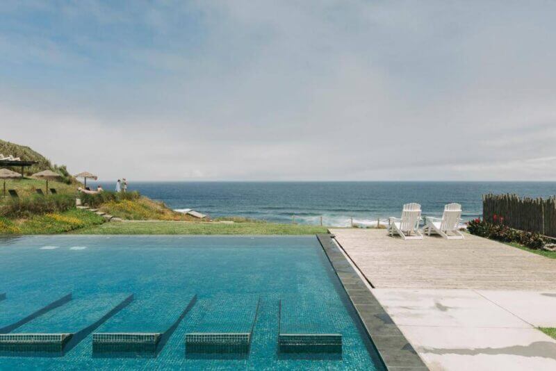 Outdoor swimming pool with submerged steps next to a wooden deck and two white chairs at Santa Barbara Resort, overlooking the grassy grounds and ocean of Sao Miguel under a partly cloudy sky.