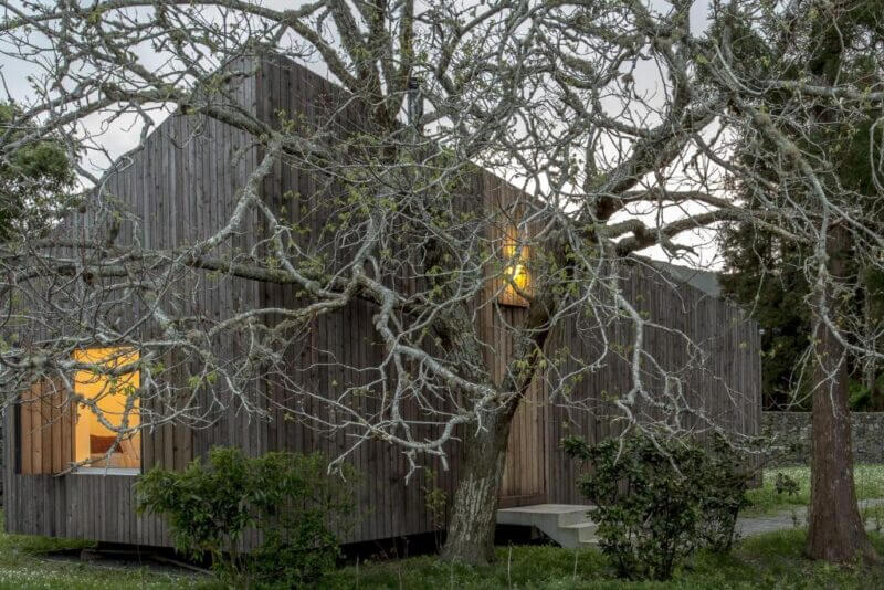 A modern wooden house with illuminated windows stands behind a leafless tree and surrounding shrubs on a cloudy day at Lake Lodge, Sete Cidades in the Azores.