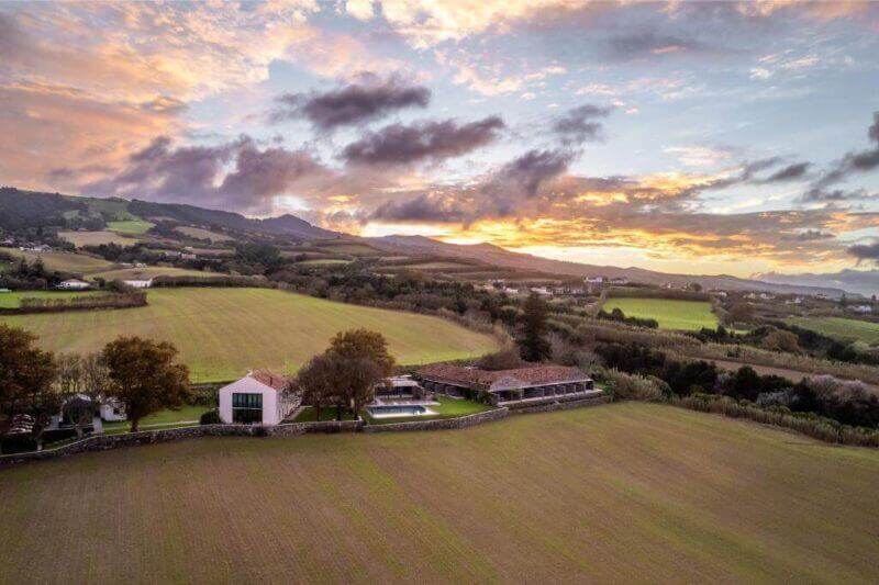 Aerial view of a countryside estate with a house, pool, and outbuildings surrounded by nature and rolling hills at sunset—your perfect Azores retreat.