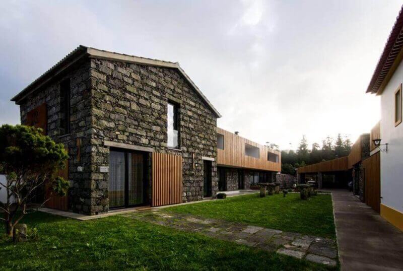 A modern building with stone and wood accents stands next to a walkway and grassy yard at Quinta Da Queiró, with another structure visible on the right side near Sete Cidades.