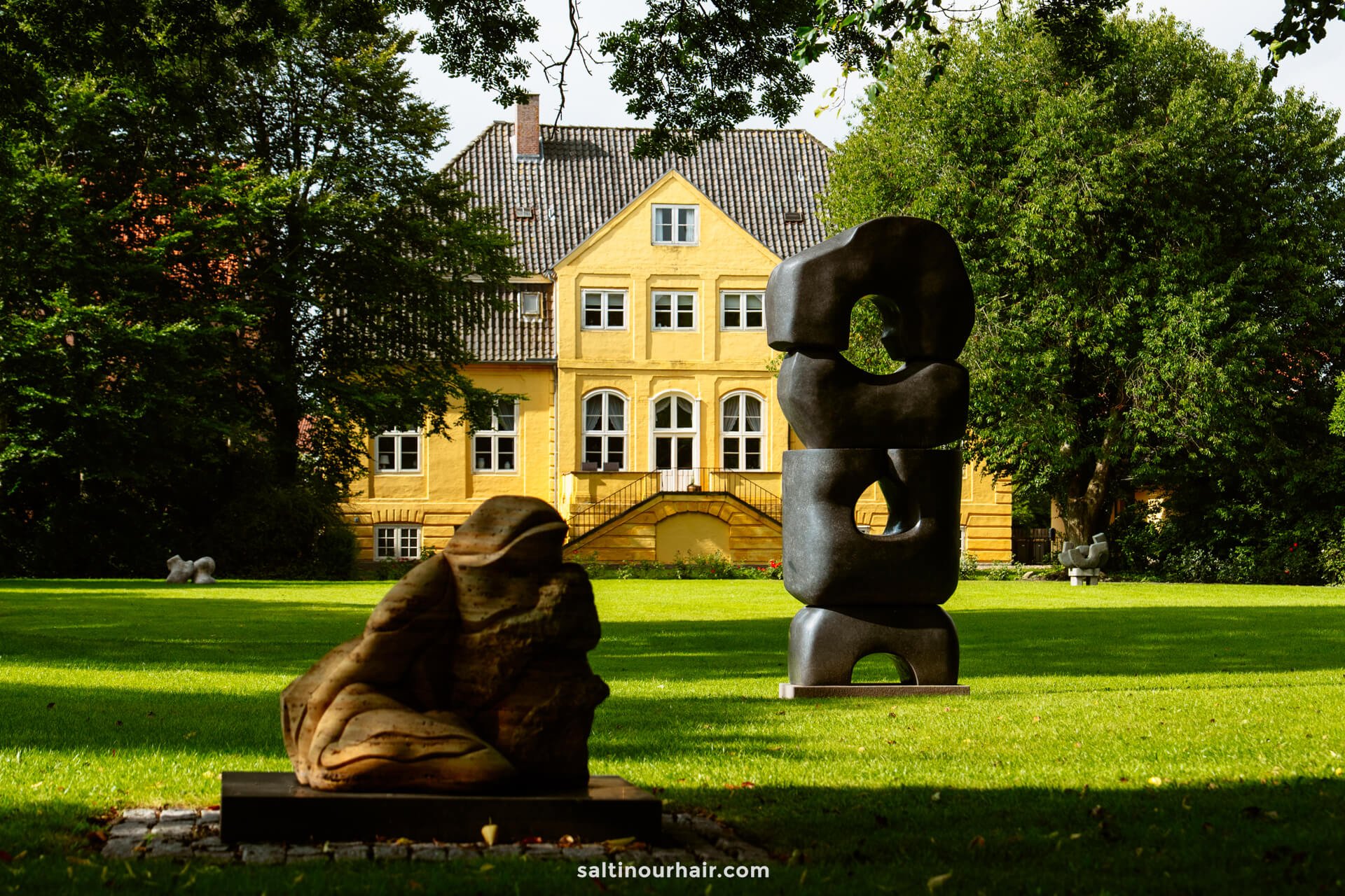 A yellow building is seen in the background with two abstract sculptures on a green lawn surrounded by trees, making it a perfect spot to add to your Denmark itinerary.