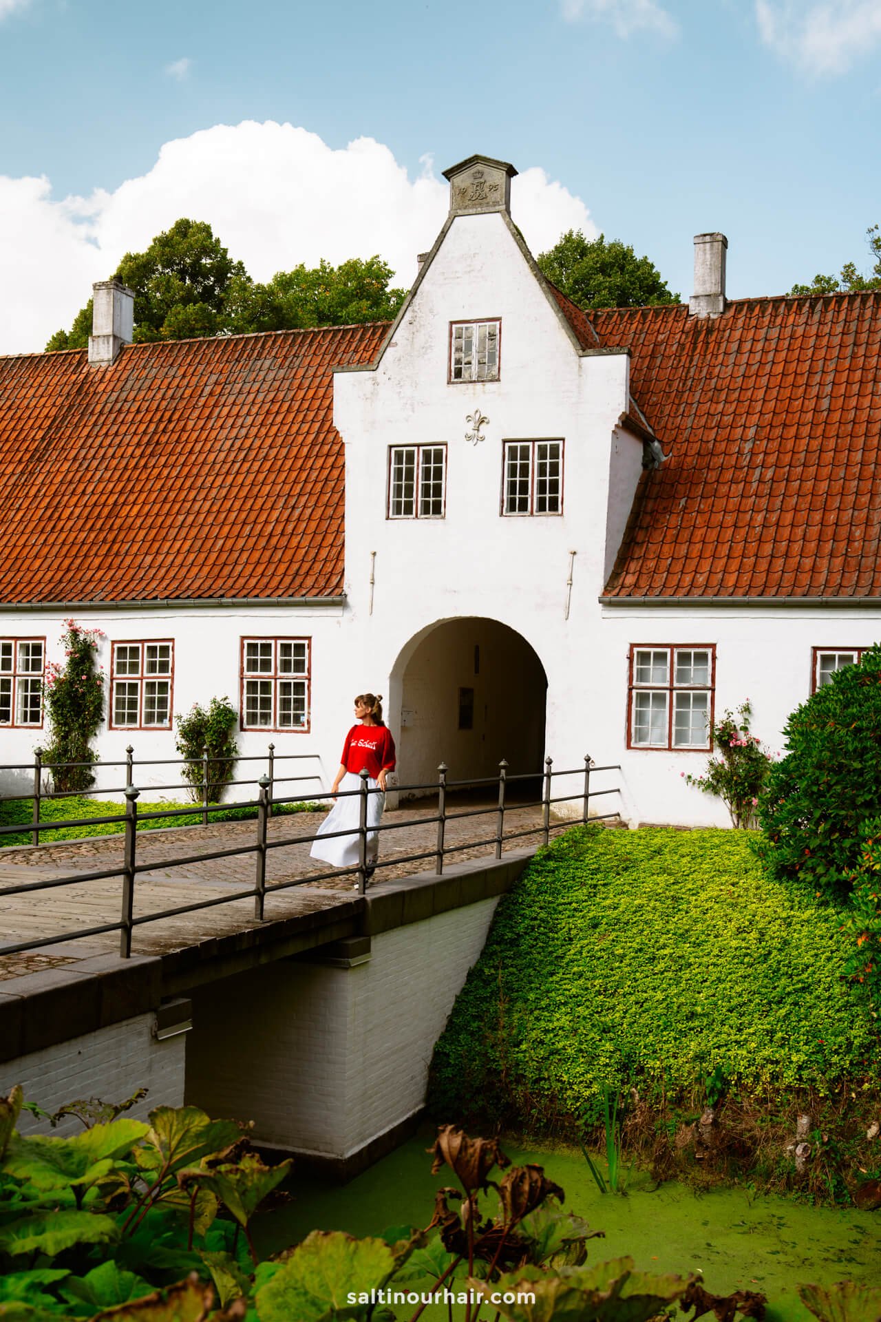 A person in a red shirt walks across a small bridge toward a white historic building with a red-tiled roof in Møgeltønder—an ideal stop on any Denmark itinerary.