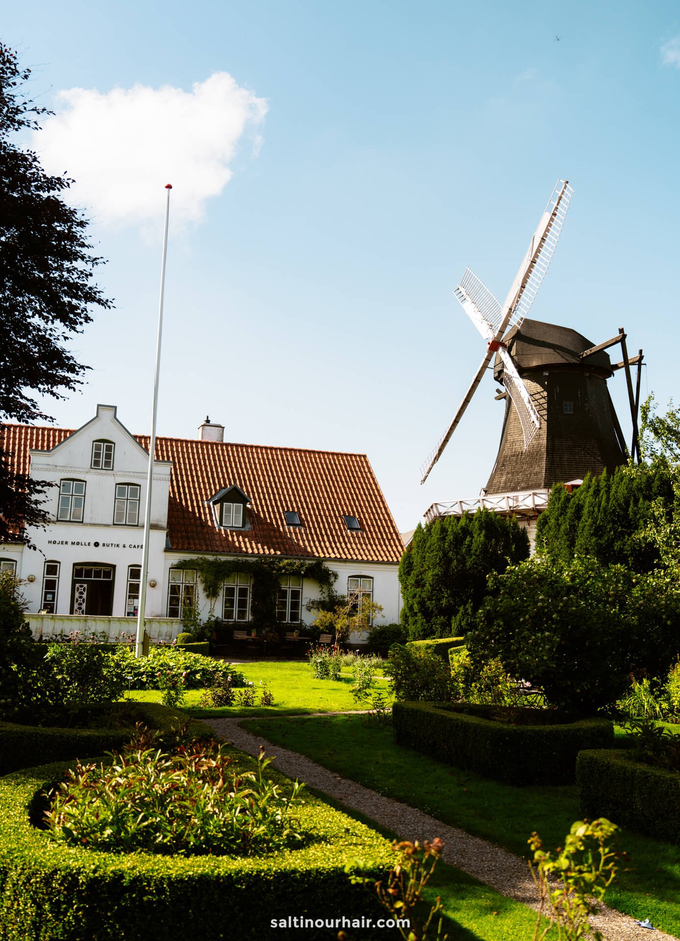 A traditional Dutch windmill stands next to a white house with a red-tiled roof in Højer, surrounded by a manicured garden under a blue sky—an idyllic scene to inspire any Denmark itinerary.