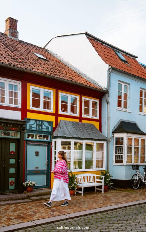 A person wearing a pink striped top and white skirt walks past colorful houses with red, yellow, and blue facades on a cobblestone street—perfect inspiration for your Denmark itinerary.