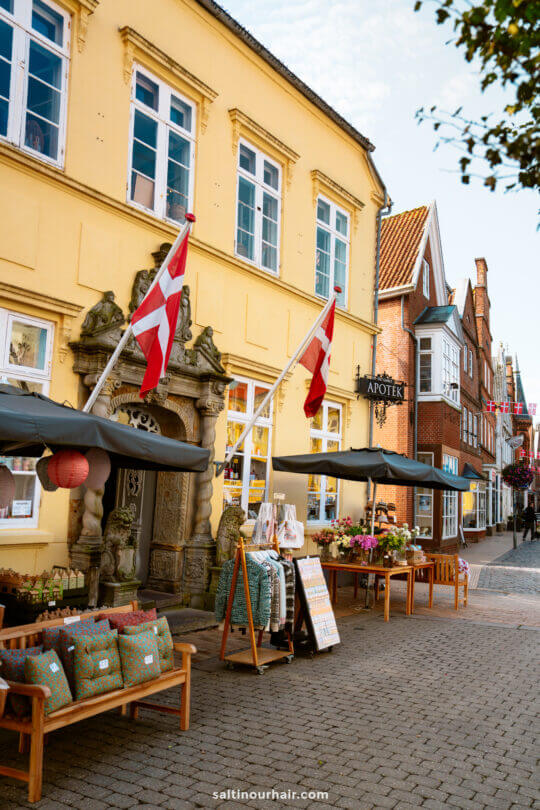 A street view of a yellow building with Danish flags in Tønder, outdoor displays of pillows and flowers, and neighboring shops—an inviting scene perfect for your Denmark itinerary in a charming European town.