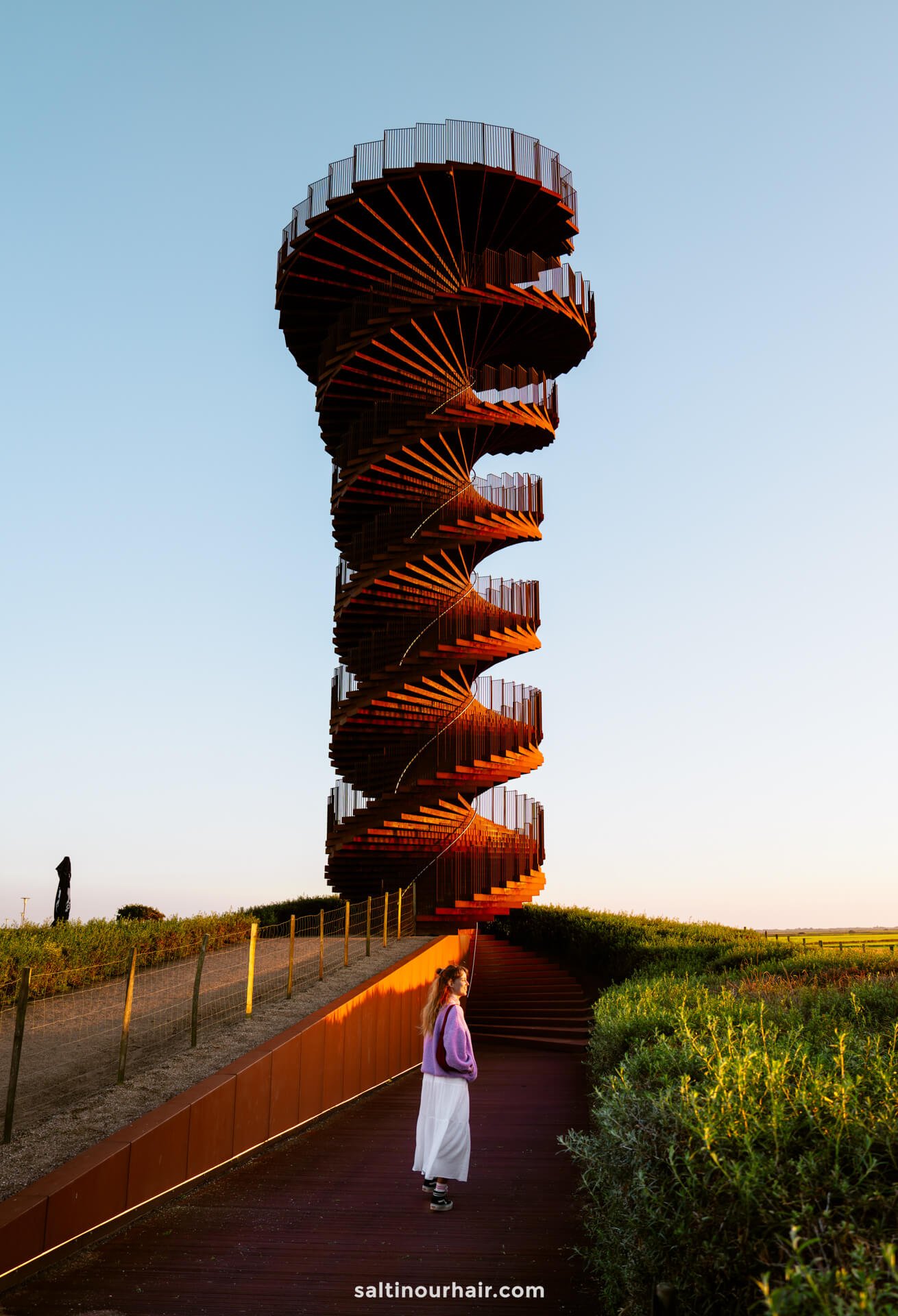 A person stands at the base of a tall, spiral observation tower called Marsk Tower at sunset—an unforgettable moment for any Denmark itinerary—as another individual takes in the view from the top, all surrounded by lush greenery and an open sky.