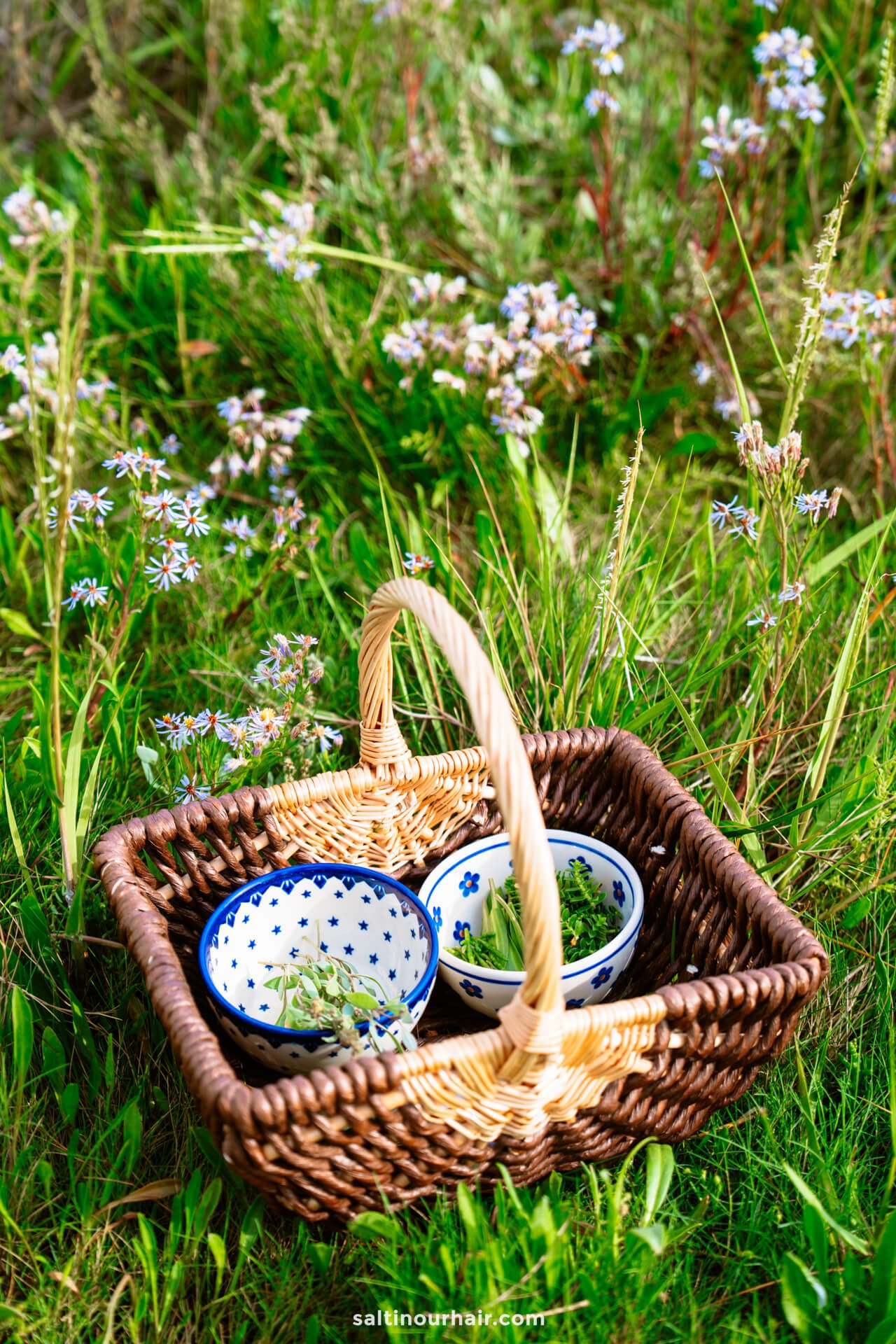 A wicker basket with three patterned bowls containing herbs, placed on grass surrounded by wildflowers—perfect inspiration for your Denmark itinerary.