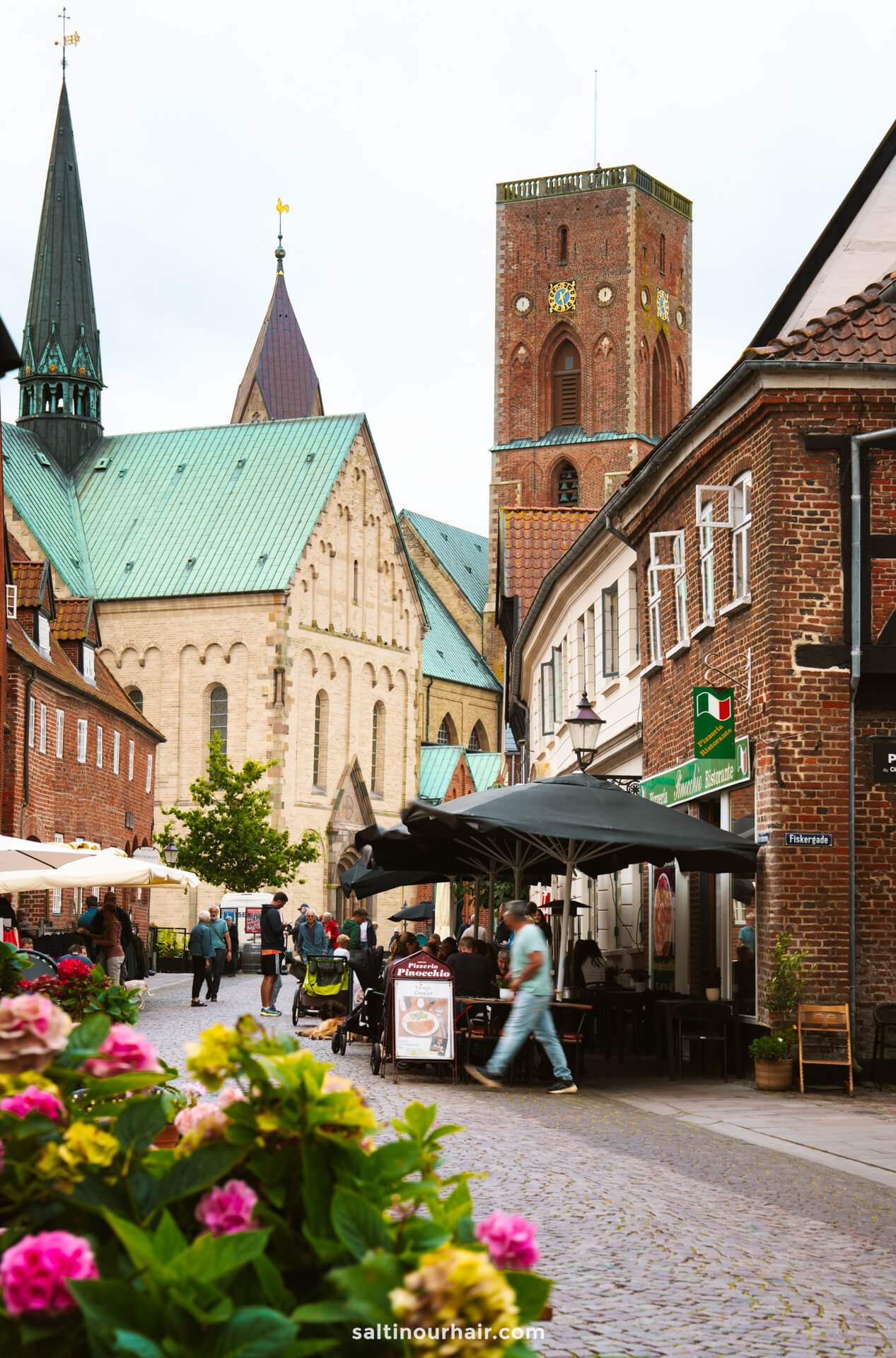 A cobblestone street with outdoor cafes and pedestrians, lined with brick buildings, leads to a large historic church with a tall tower and green roof in Ribe—an ideal sight on any Denmark itinerary.