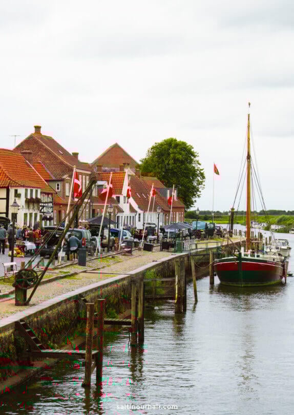 A docked sailboat on a canal beside a walkway lined with red-roofed buildings in Ribe, with several red-and-white flags. People are gathered along the scenic waterfront.