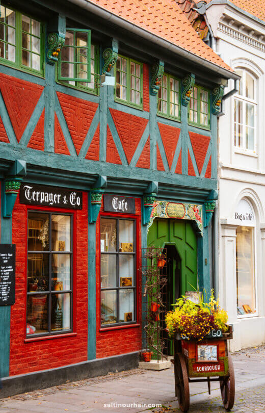 A traditional half-timbered building with teal and red accents houses a café and shop in Ribe; perfect for your Denmark itinerary, a cart with flowers and a “sourdough” sign stands outside the entrance.