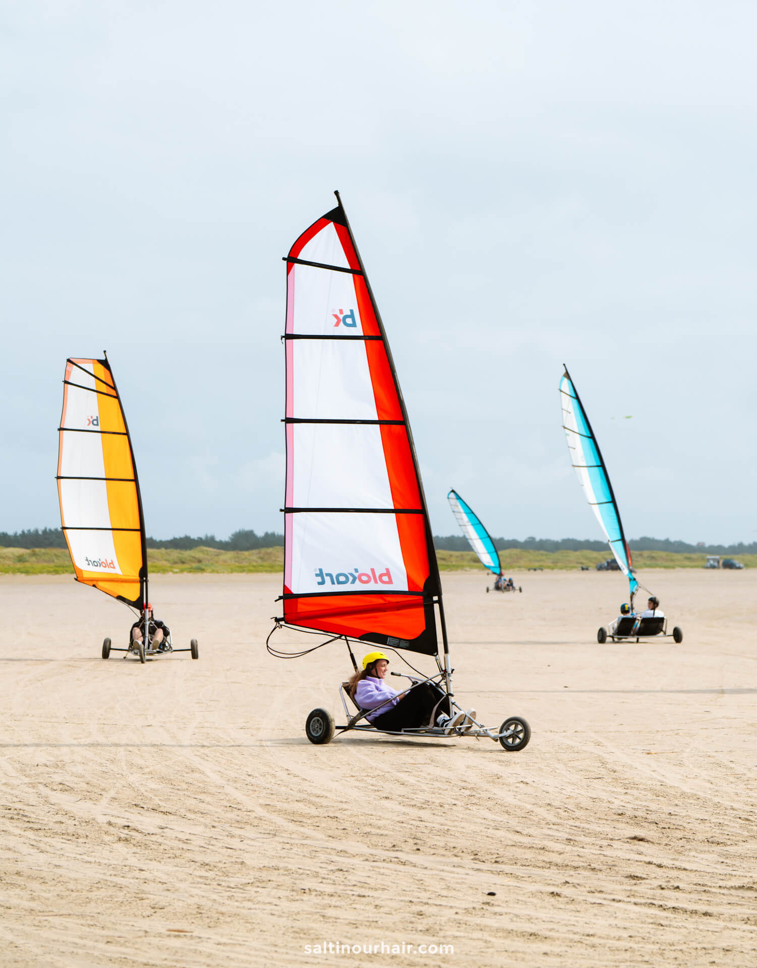Three people ride blokarts with colorful sails on a sandy beach under a cloudy sky, with grassy dunes in the background—a perfect addition to your Denmark itinerary.