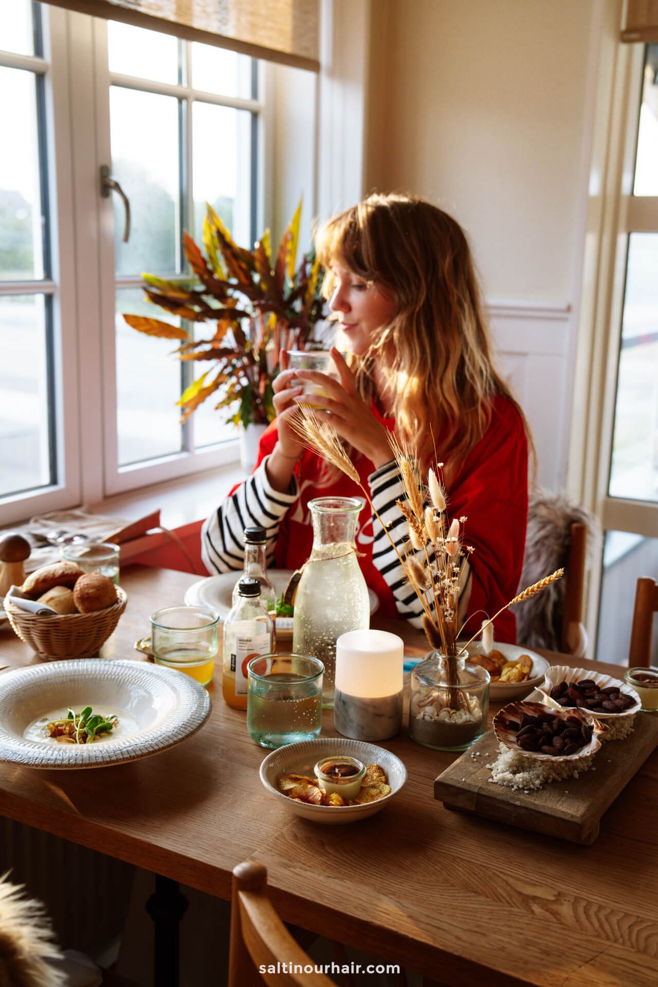 A woman sits at a wooden table set with food, drinks, and candles near a window, holding a glass and looking outside—perhaps reminiscing about her recent Denmark itinerary.