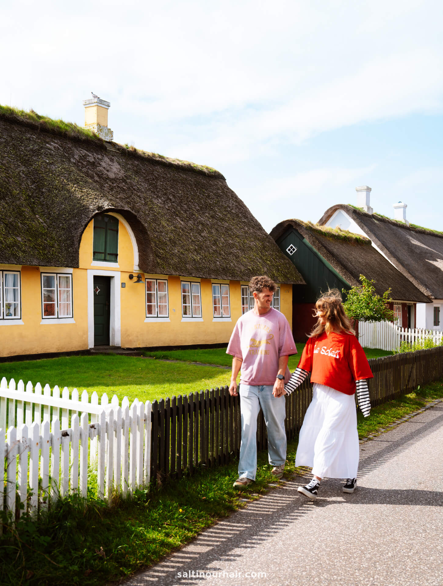 Two people walk on a road beside yellow houses with thatched roofs, white picket fences, and green lawns on a sunny day in Sønderho—an idyllic scene perfect for any Denmark itinerary.