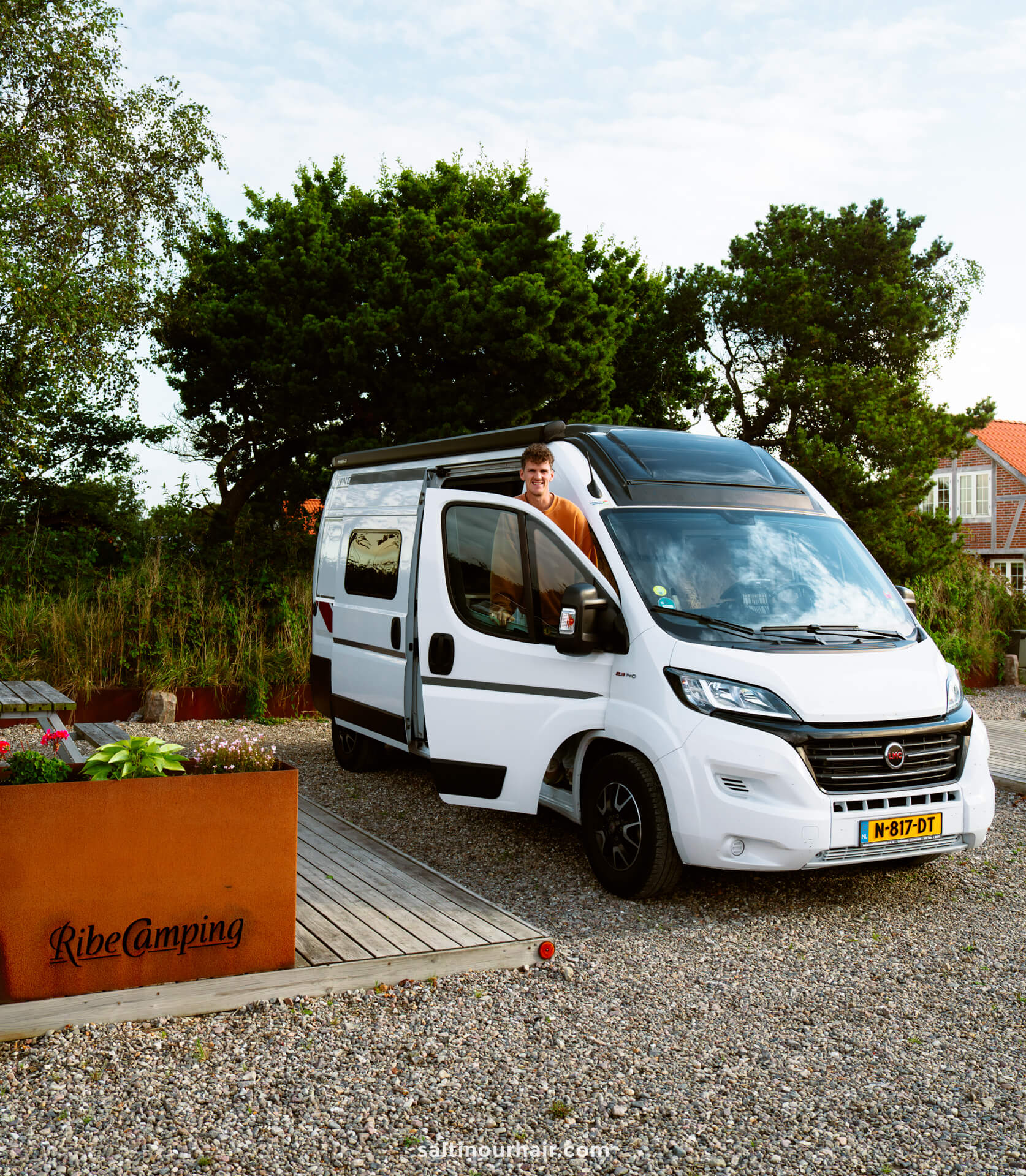 A person stands in the open side door of a white camper van parked at Ribe Camping, Denmark—a perfect stop on any Denmark road trip—with trees and buildings in the background.