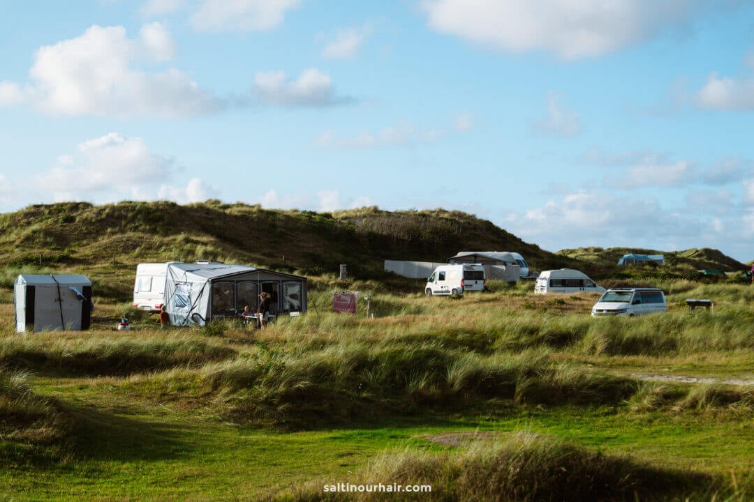 Several camper vans, tents, and cars are parked among grassy sand dunes under a partly cloudy sky on a camping site—an ideal stop on any Denmark itinerary.