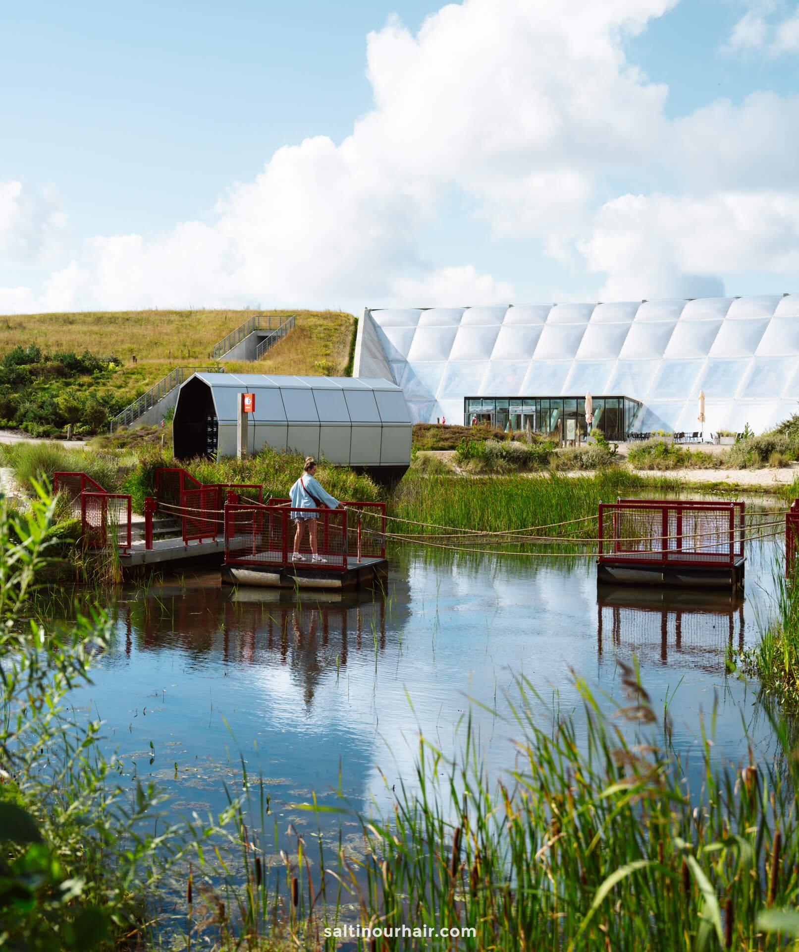 A person stands on a red footbridge over a pond with reeds at Naturkraft Outdoor Park Denmark, near modern geometric buildings and grassy hills under a partly cloudy sky.
