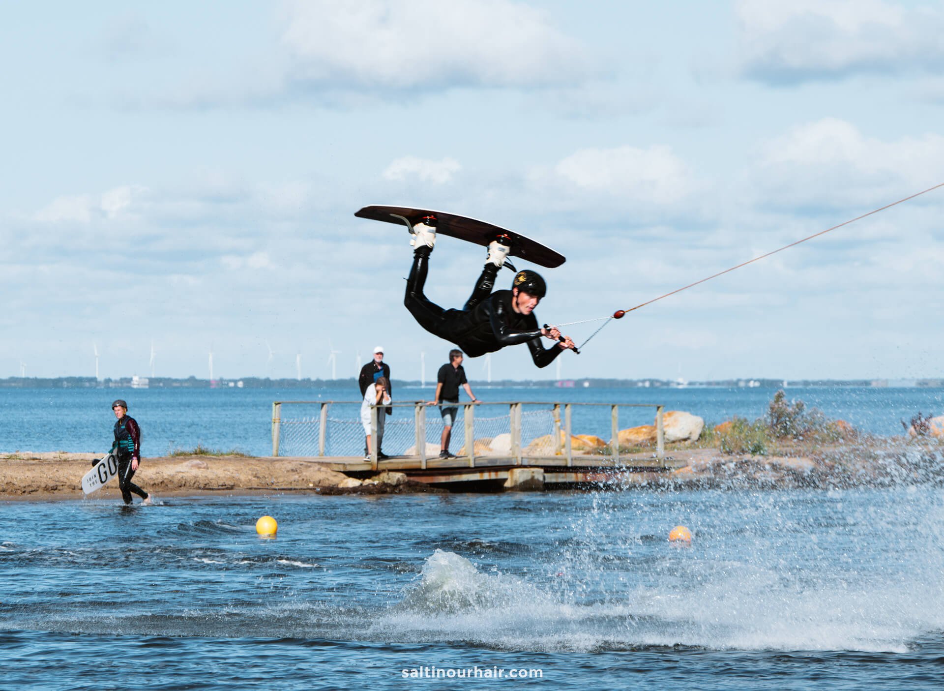 A person in a wetsuit performs an airborne trick on a wakeboard, holding a tow rope, while spectators—perhaps planning their Denmark itinerary—watch from the water’s edge.