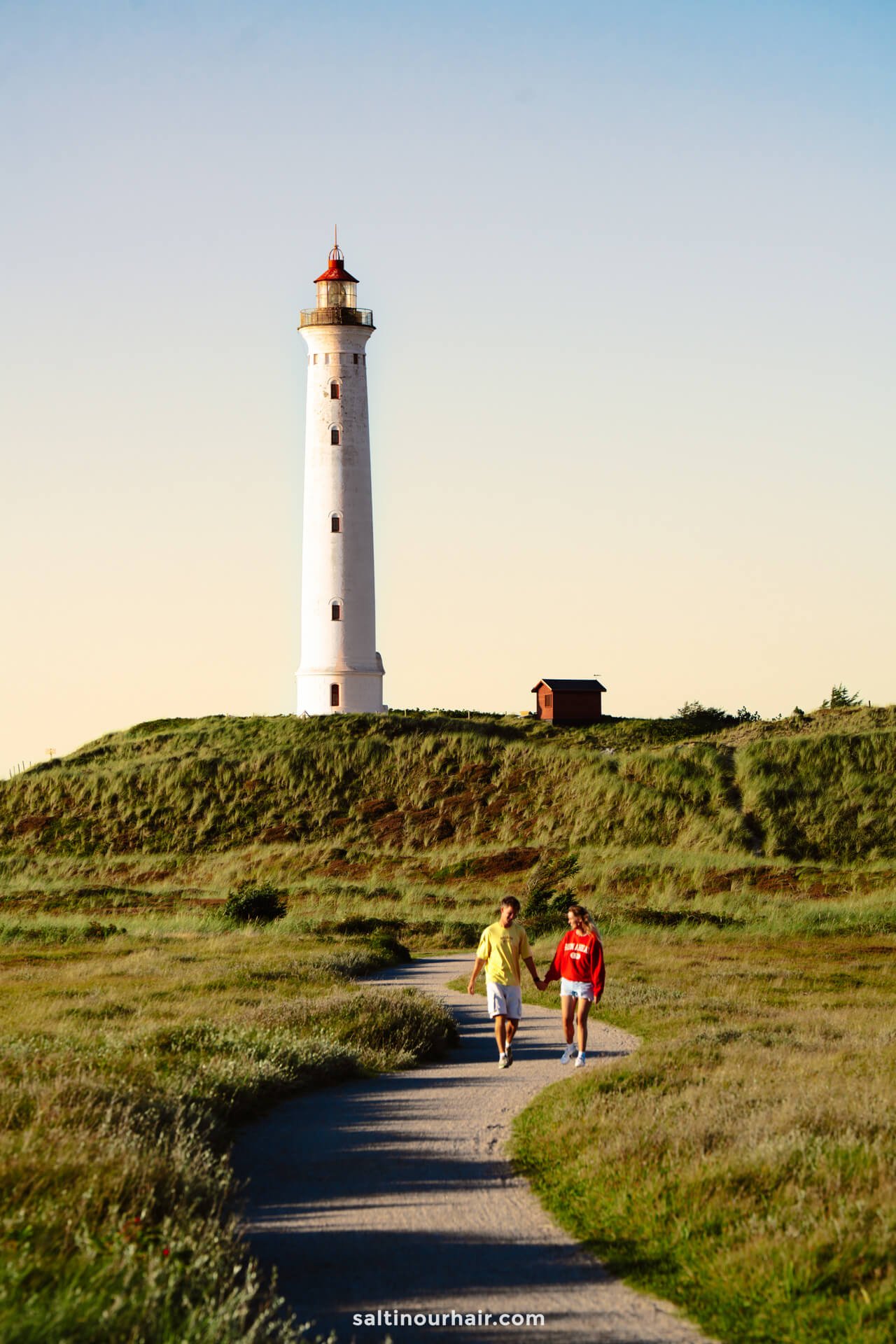 Two people walk on a path through grassy fields toward Lyngvig Fyr, Denmark’s tall white lighthouse with a red top, under a clear sky.