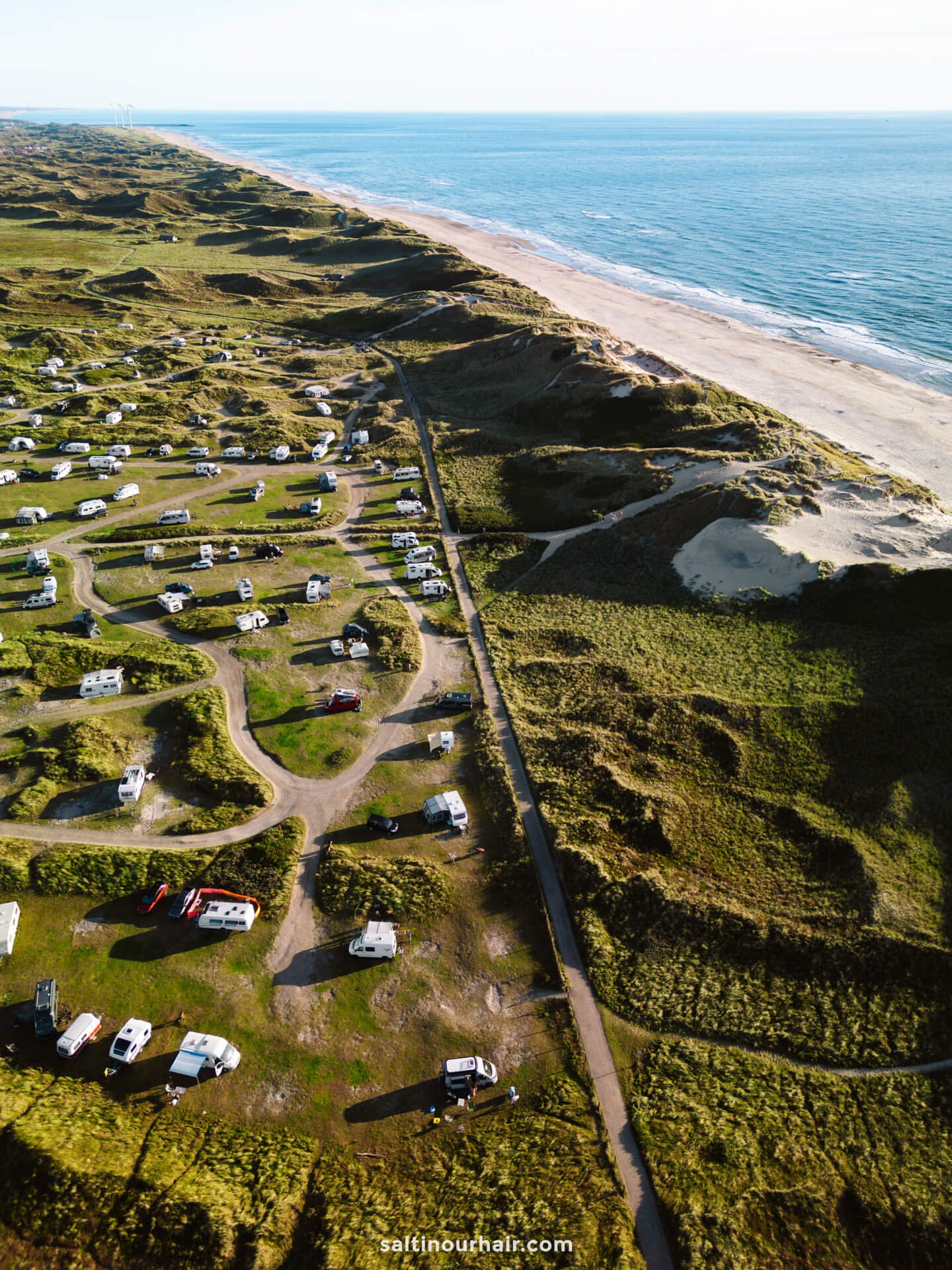 Aerial view of a coastal campsite, perfect for any Denmark itinerary, with numerous RVs and winding roads near dunes and a sandy beach beside the ocean.