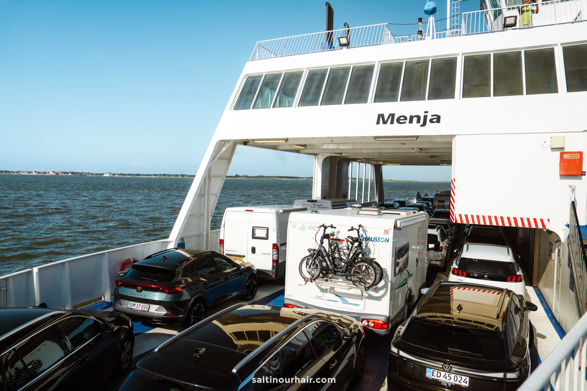 Cars and a camper van with bicycles on the back are parked on the open deck of a ferry to Fanø island, an ideal sight for any Denmark itinerary, as it travels across serene waters under a clear sky.