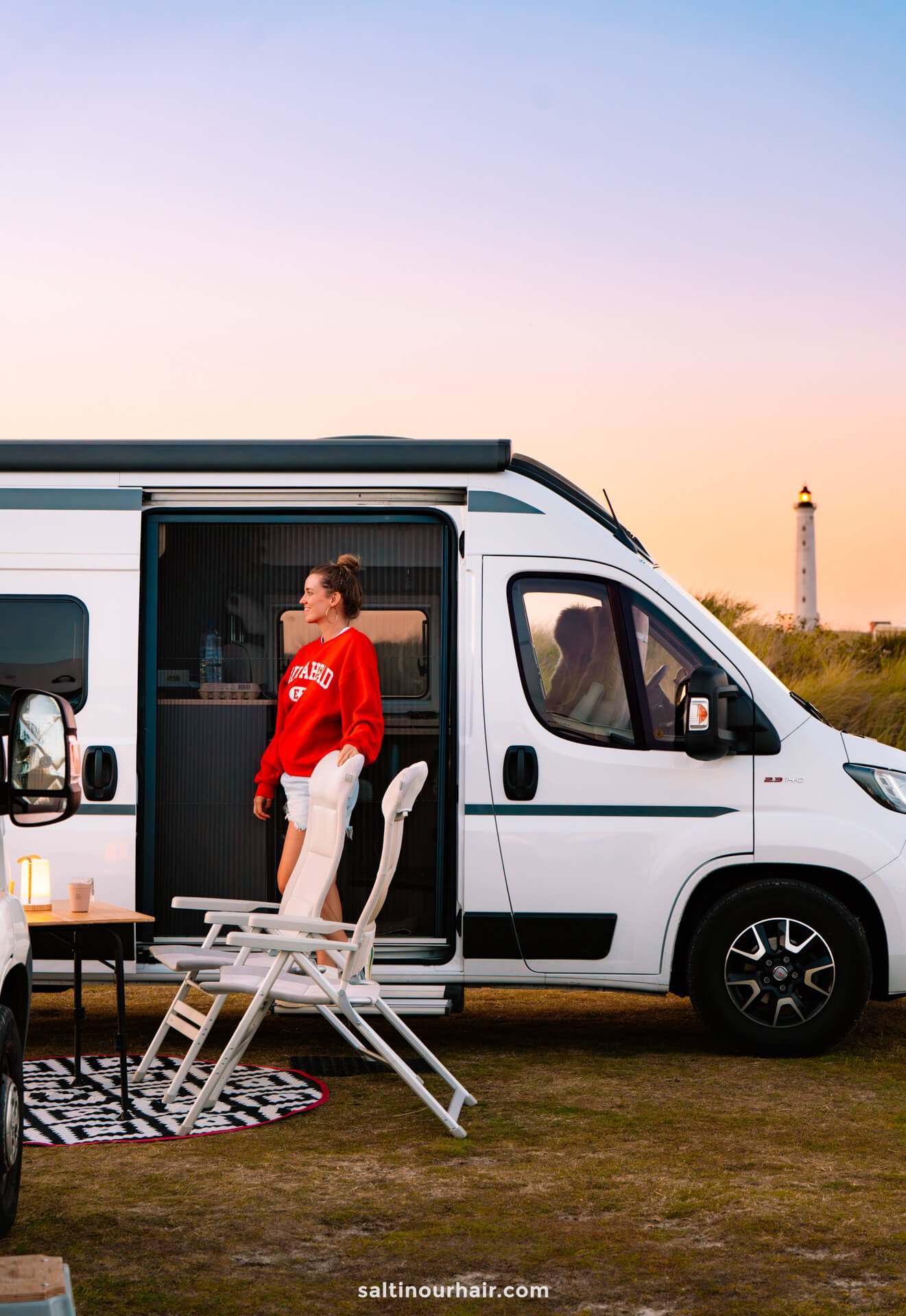 A woman stands in the doorway of a white camper van parked on grass at sunset, with a chair, table, and Lyngvig Fyr lighthouse in the background—an idyllic stop on any Denmark road trip.