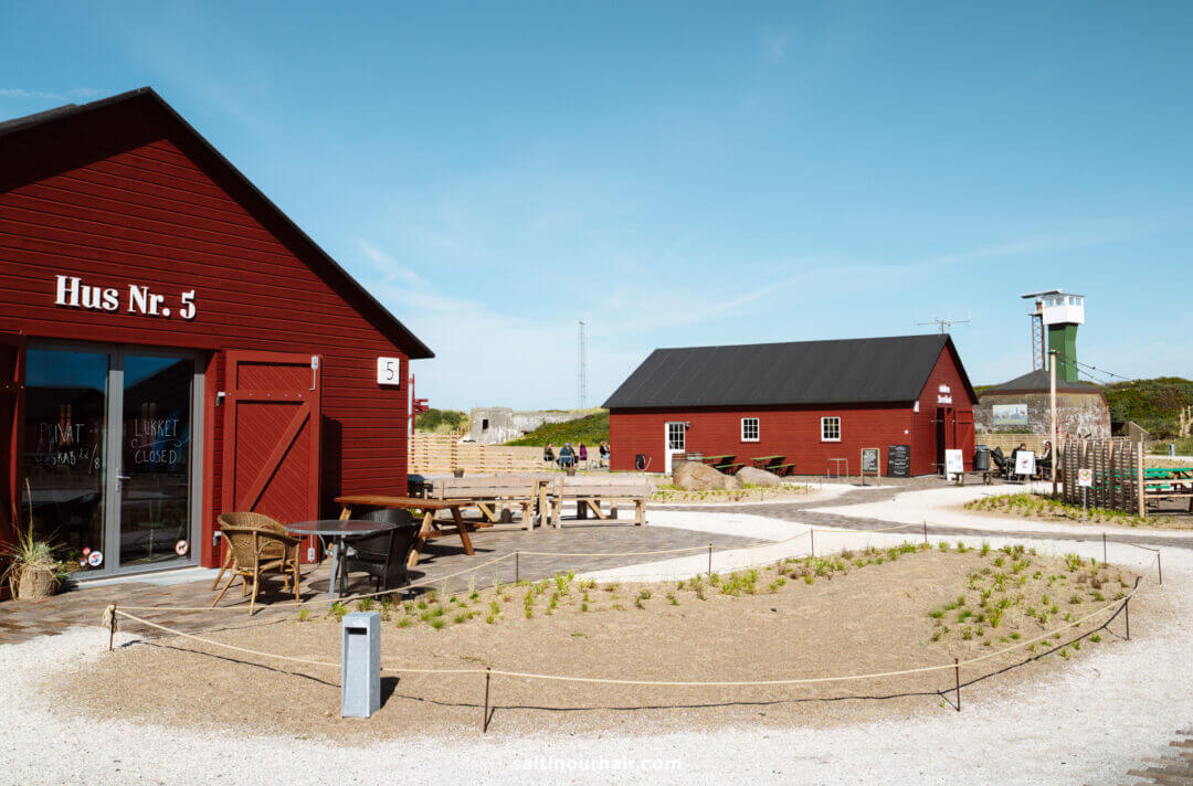 Scene with red wooden buildings, outdoor seating, gravel paths, and sparse landscaping under a clear blue sky—a charming stop to include on your Denmark itinerary. A watchtower rises in the background.