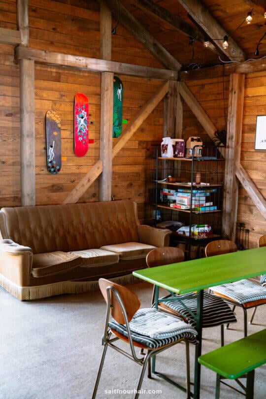 A cozy room with a brown sofa, green tables, wooden chairs, and a metal shelf of board games—perfect for planning your Denmark itinerary among skateboards mounted on wooden walls.