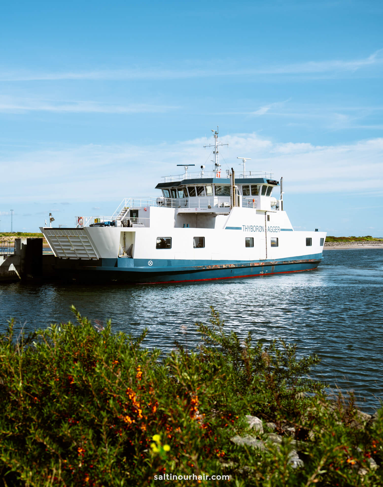 A white and blue ferry named Thyborøn-Agger is docked near the shore with greenery and water in the foreground under a clear sky, making it a scenic stop on any Denmark itinerary.