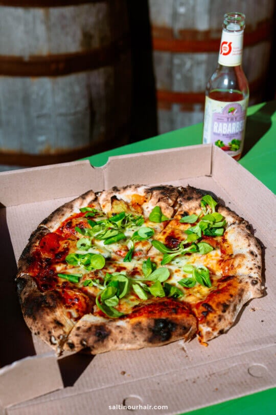 A wood-fired pizza topped with green leaves sits in an open cardboard box on a green table next to a bottle of rhubarb soda—perfect for your Denmark itinerary. Two wooden barrels are visible in the background.