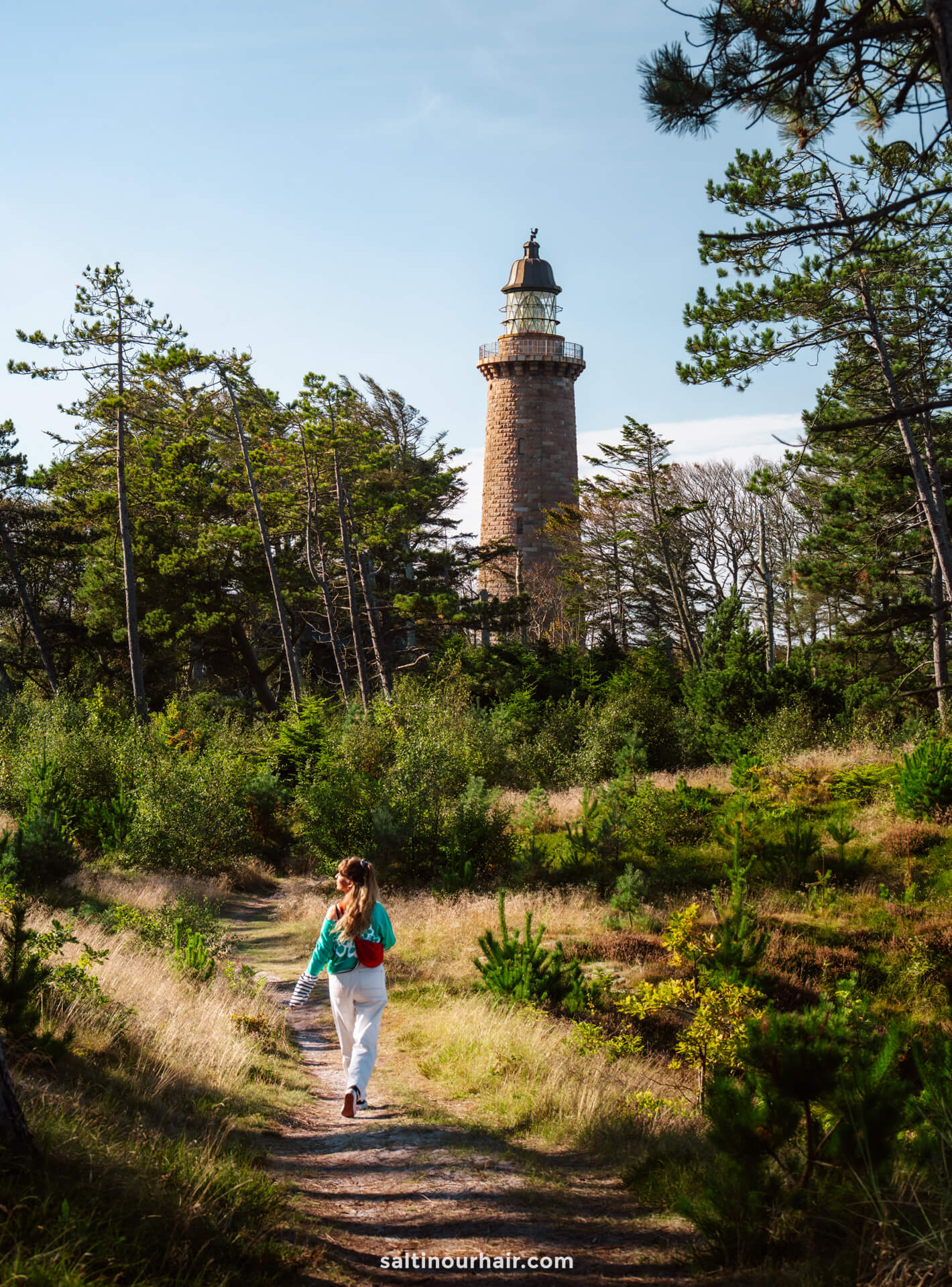 A person walks along a forest path toward the tall brick lighthouse Lodbjerg Fyr surrounded by trees on a sunny day—a perfect stop on any Denmark itinerary.