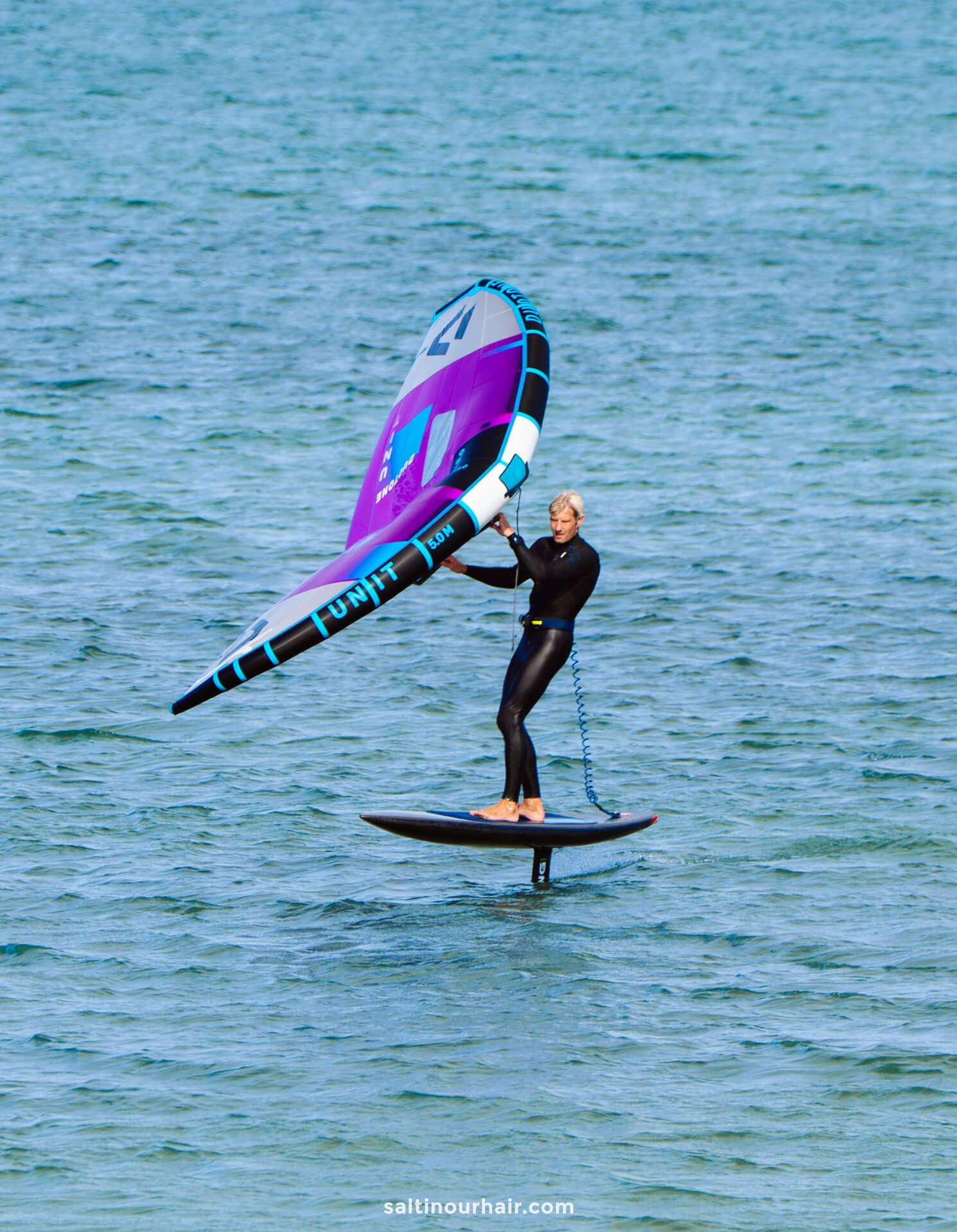 A person wearing a wetsuit rides a hydrofoil board on the water, holding a purple and blue handheld sail—an adventure you might add to your Denmark itinerary.