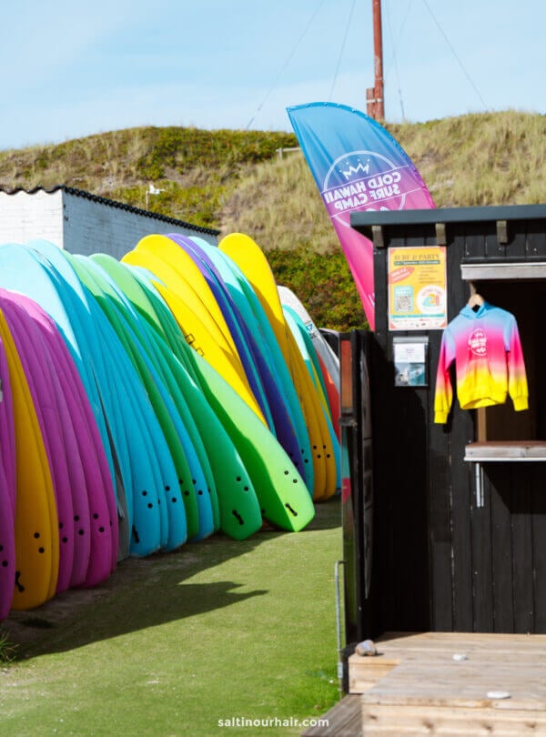 Colorful surfboards are lined up next to a black wooden hut with a rainbow sweatshirt hanging outside—an inviting stop on any Denmark itinerary, with a flag and grassy sand dunes visible in the background.