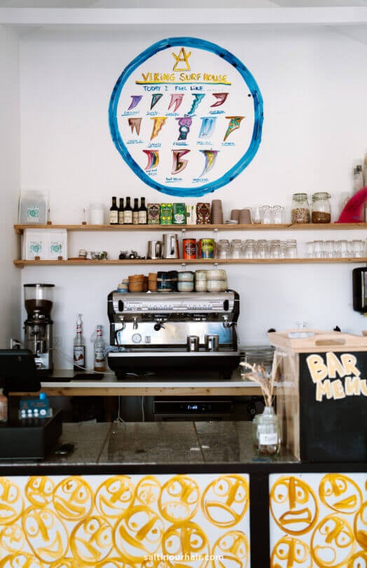 A coffee bar with a large espresso machine, shelves of cups and jars, and a colorful surf-themed menu displayed on the wall above—an inviting stop to add to your Denmark itinerary.