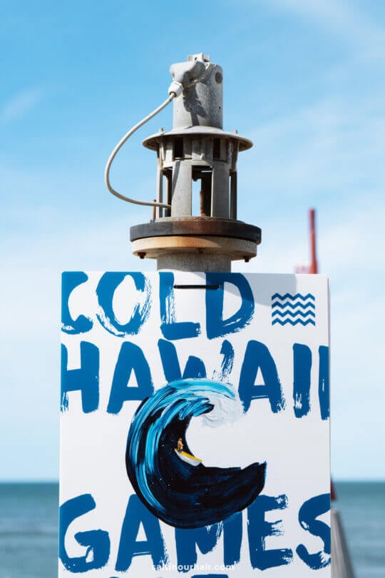 A sign reading Cold Hawaii Games with a painted wave sits beneath mechanical equipment, set against a blue sky and ocean—an ideal stop on any Denmark itinerary.