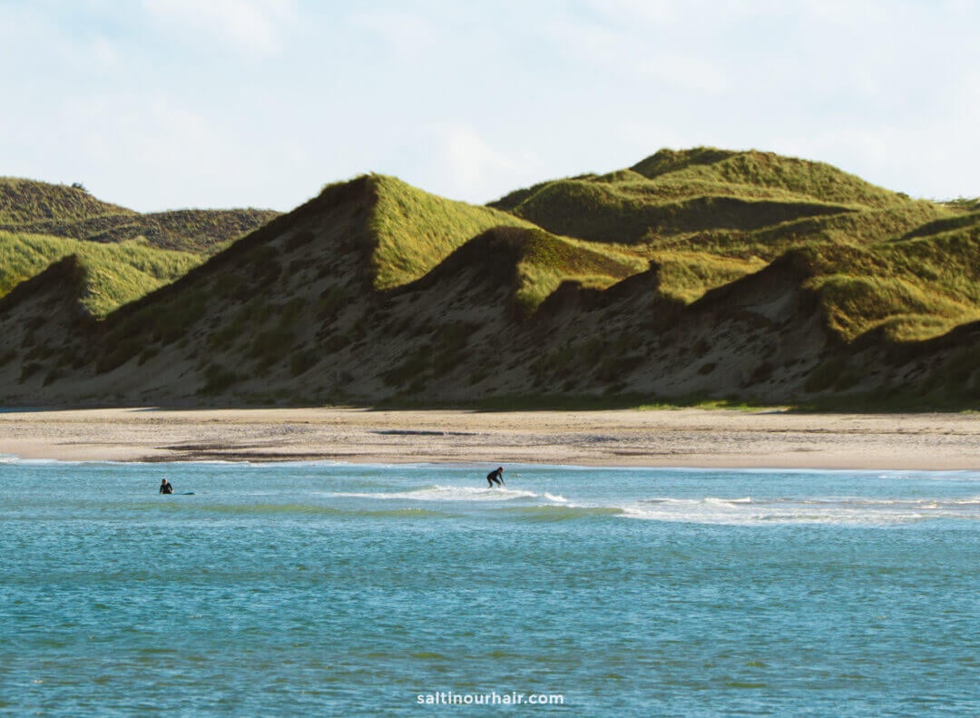 Two people surf small waves near a sandy beach with grassy, sloping dunes in the background under a partly cloudy sky—a perfect scene to include in your Denmark itinerary.