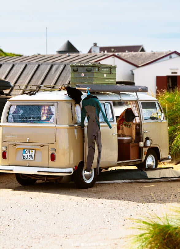 A beige camper van with surf gear, including a wetsuit hanging on the side, parked near beach houses on a sunny day—perfect for any Denmark itinerary.