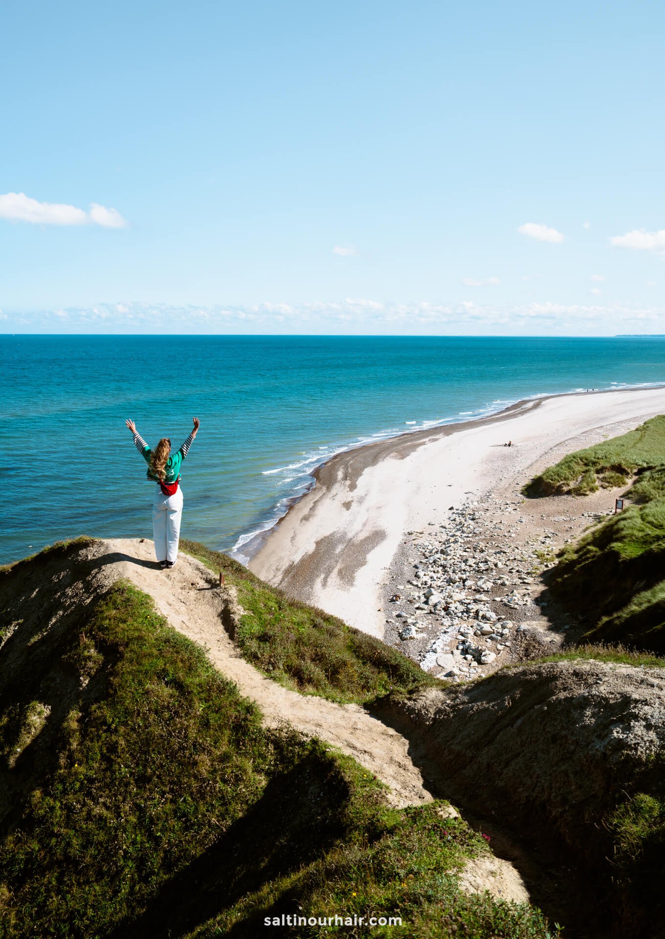 A person stands on a grassy cliff called Bulbjerg Klint overlooking a sandy beach and blue ocean under a clear sky, arms raised in the air—an inspiring stop on any Denmark itinerary.
