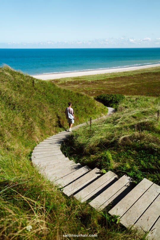 A person walks down a wooden boardwalk through grassy dunes toward a sandy beach and blue sea at Bulbjerg Klint, capturing the serene beauty perfect for any Denmark itinerary.