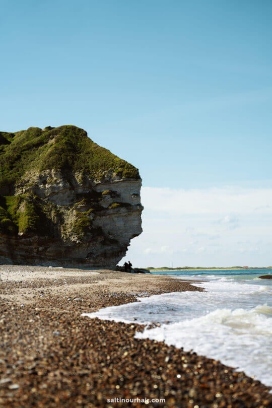 Bulbjerg Klint cliff covered in greenery rises beside a pebble beach, making it a perfect stop on any Denmark itinerary, with waves gently reaching the shore under a clear blue sky.