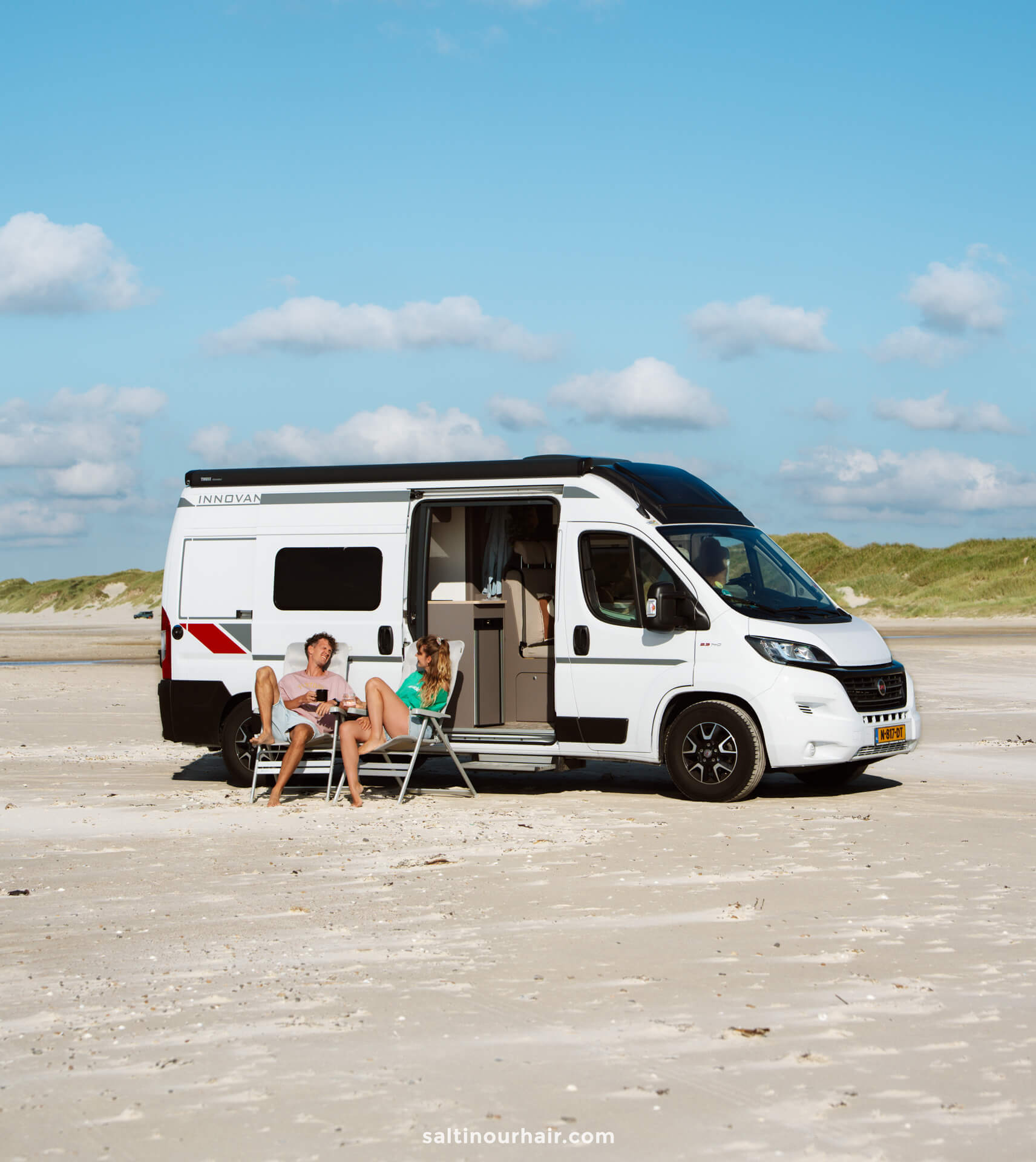 Two people sit on chairs outside a white camper van parked on a sandy beach, enjoying a peaceful stop along their Denmark road trip itinerary, with grassy dunes in the background under a blue sky with clouds.