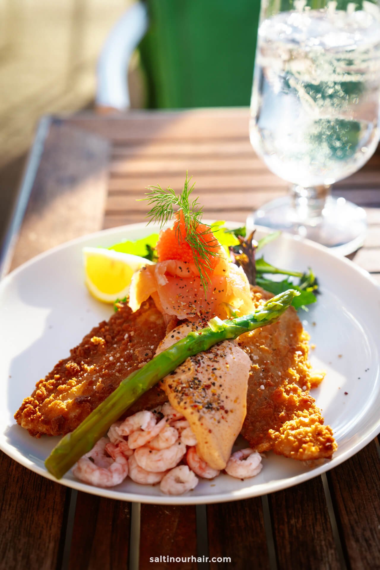 A plate of breaded fish fillets topped with smoked salmon, shrimp, a lemon wedge, asparagus, herbs, and black pepper—this Danish-inspired meal could be the highlight of your Denmark itinerary, enjoyed with a glass of water on a wooden table.