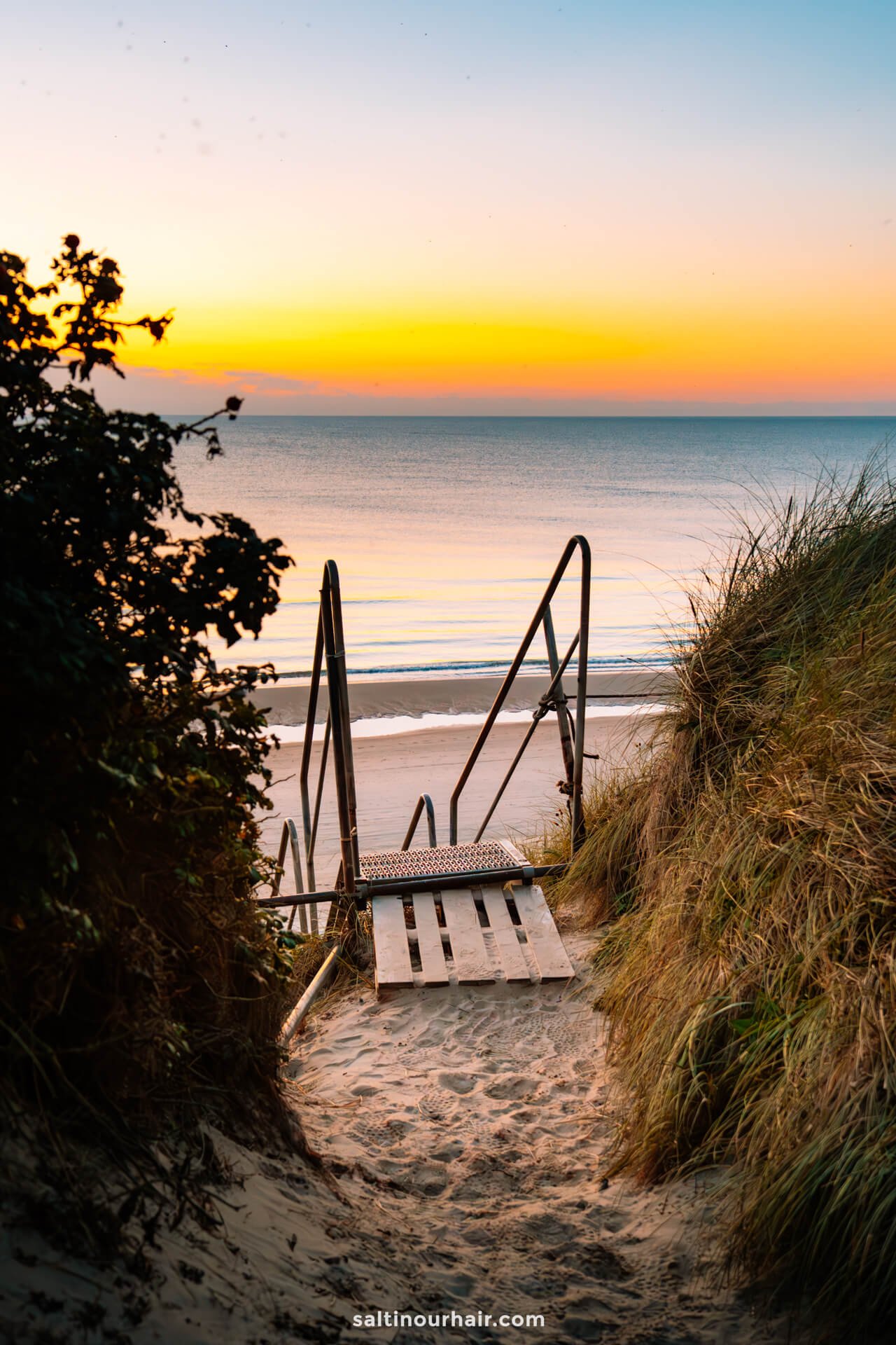 A metal staircase leads down sandy dunes to a calm beach at sunset, with orange and blue hues in the sky and gentle waves—an ideal stop on any Denmark itinerary.